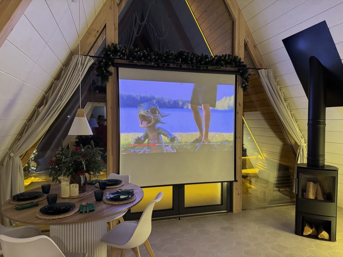 Домашний кинотеатр в A-frame: проектор с мультфильмом на стене, обеденный стол с сервировкой, чёрная дровяная печь и гирлянды